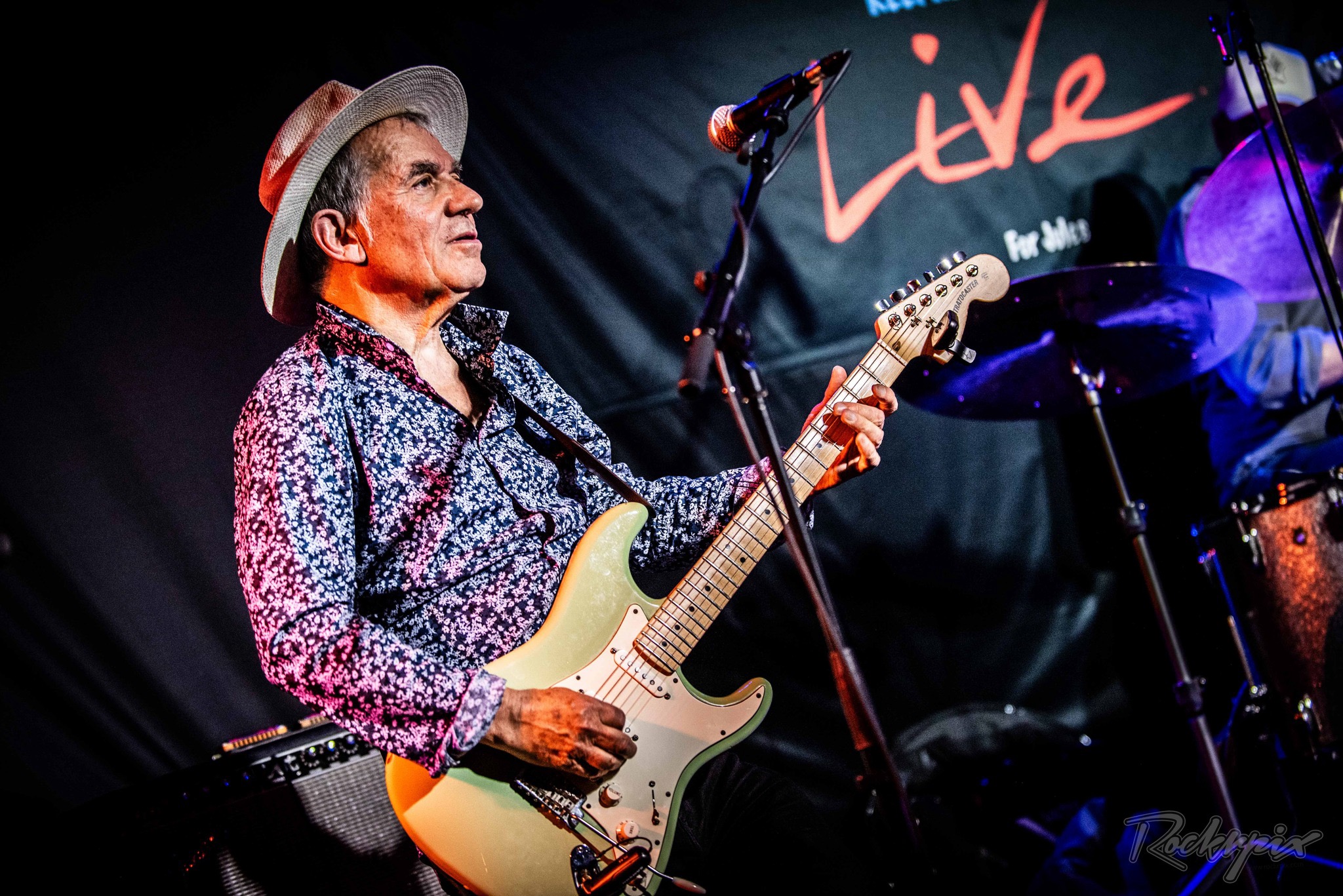 Robin Bibi in a white hat playing his Stratocaster under blue stage lighting, gazing upward