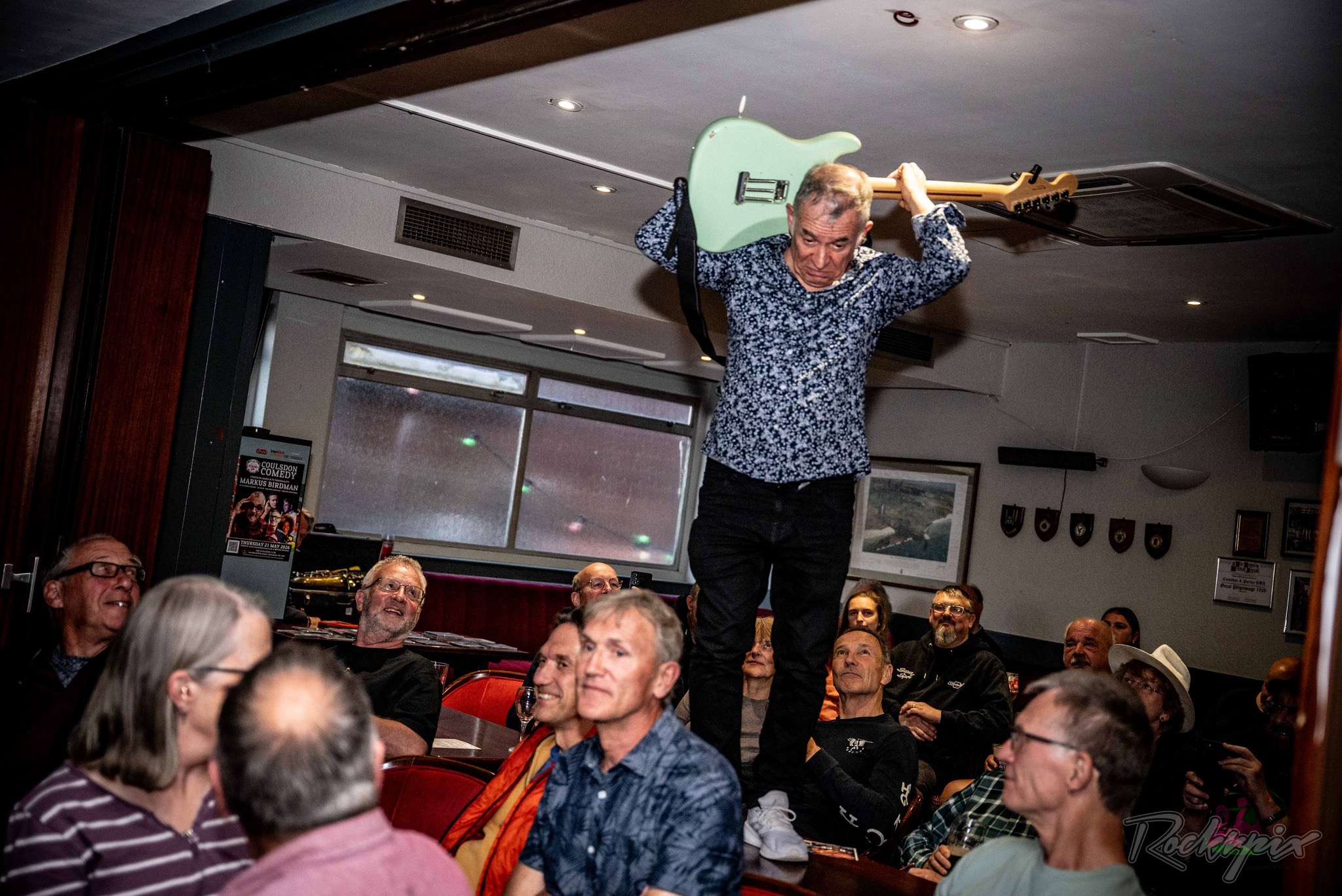 Robin Bibi standing on a table holding his guitar above his head over a seated, smiling audience