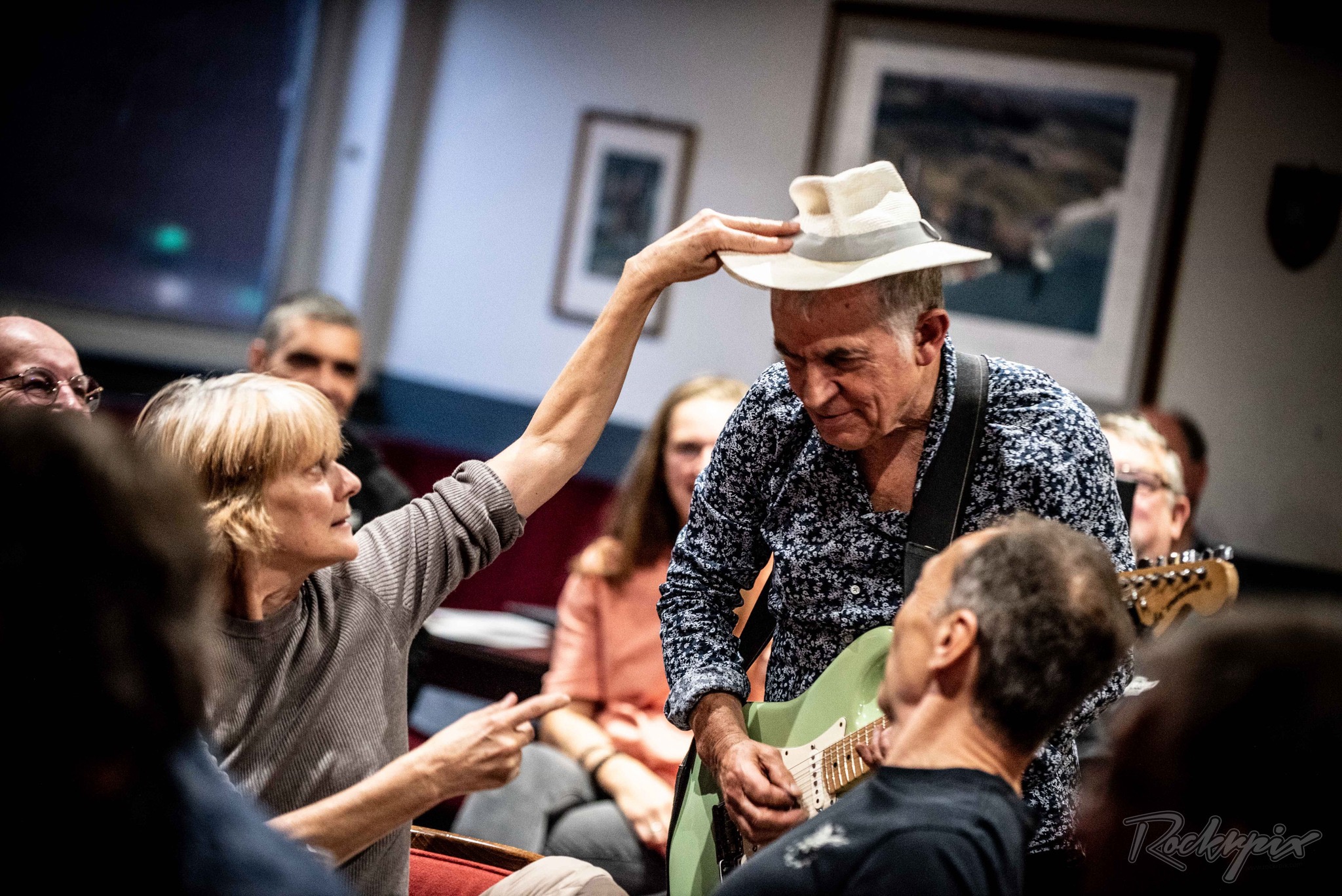 A woman placing Robin Bibi's white hat on his head as he plays guitar among the audience